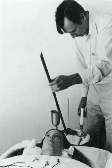 Un uomo in camicia bianca regge un’asta sopra una donna sdraiata con elettrodi sul petto; sul tavolo un calice, una candela e un libro. Foto d’archivio