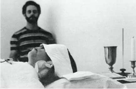 Una donna distesa con il capo coperto da un panno durante il channeling, un uomo barbuto sullo sfondo, un calice e una candela accanto. Foto d’archivio