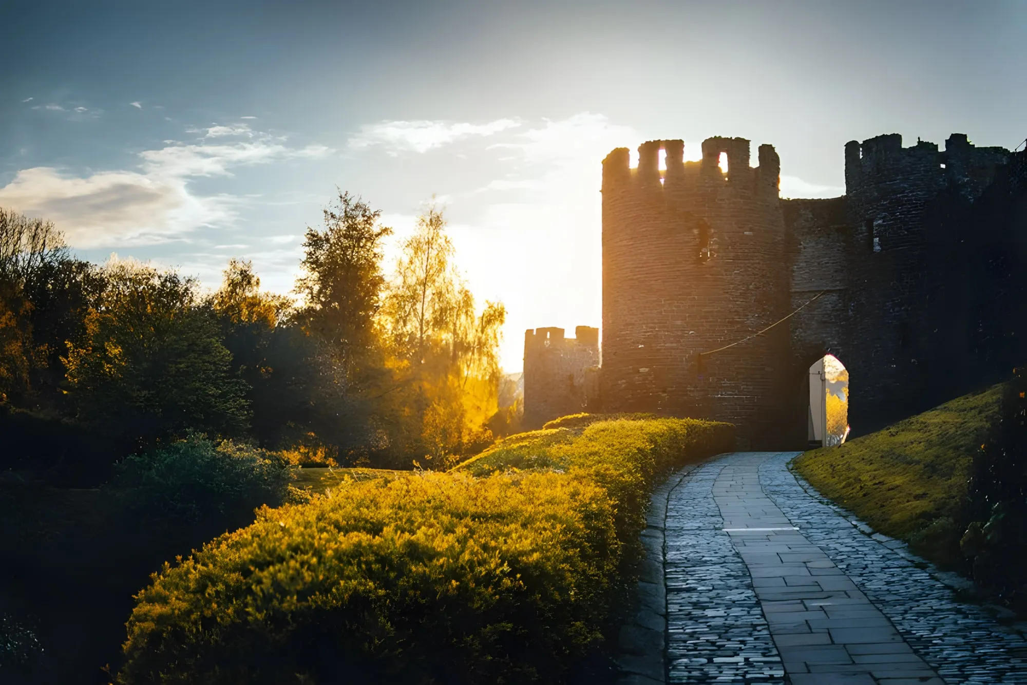 Torre di un castello in pietra illuminata dal sole al tramonto, con un sentiero lastricato che porta al portone