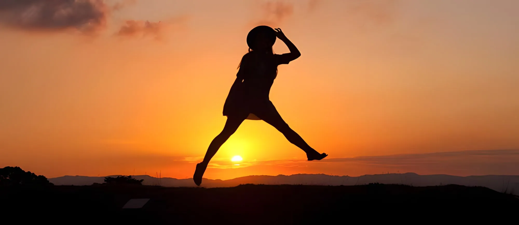 Silhouette di una donna con il cappello che salta in aria davanti a un tramonto arancione