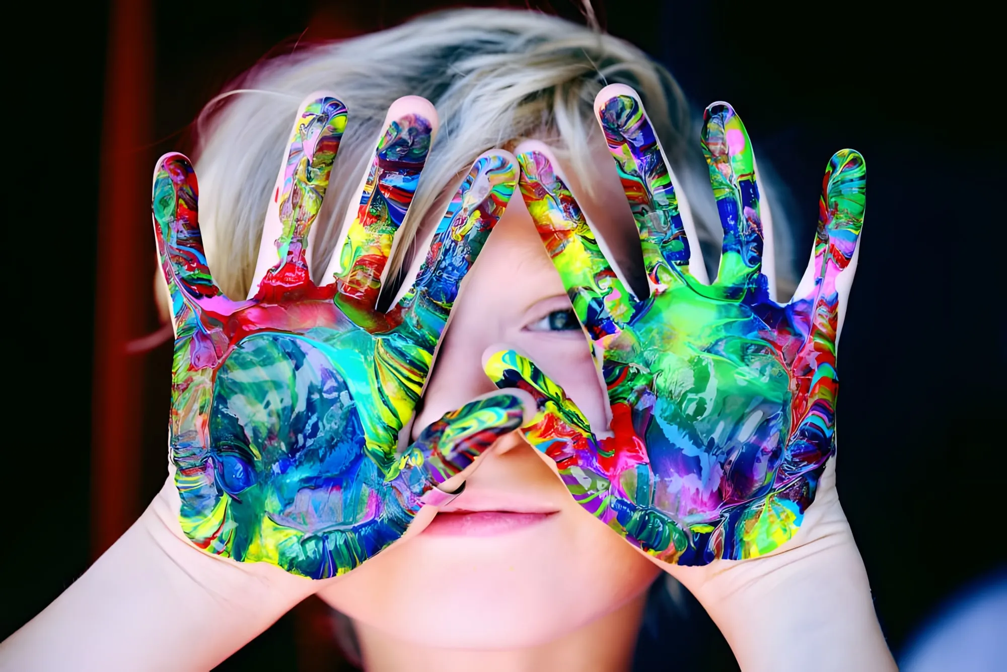 Bambino con le mani dipinte di colori vivaci che gli coprono in parte il viso sorridente