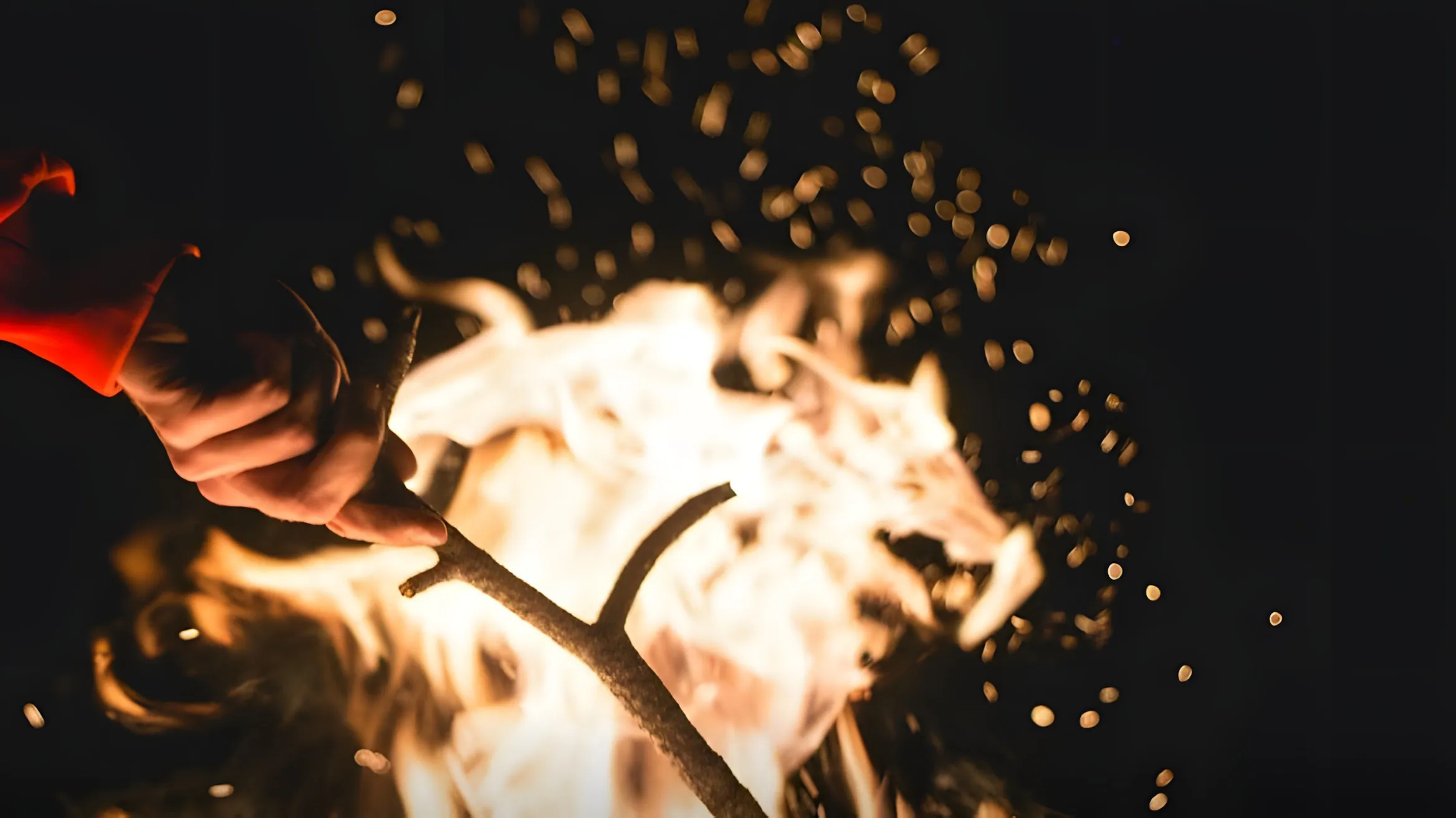 Una mano attizza con un ramo un falò nella notte, tra fiamme e scintille