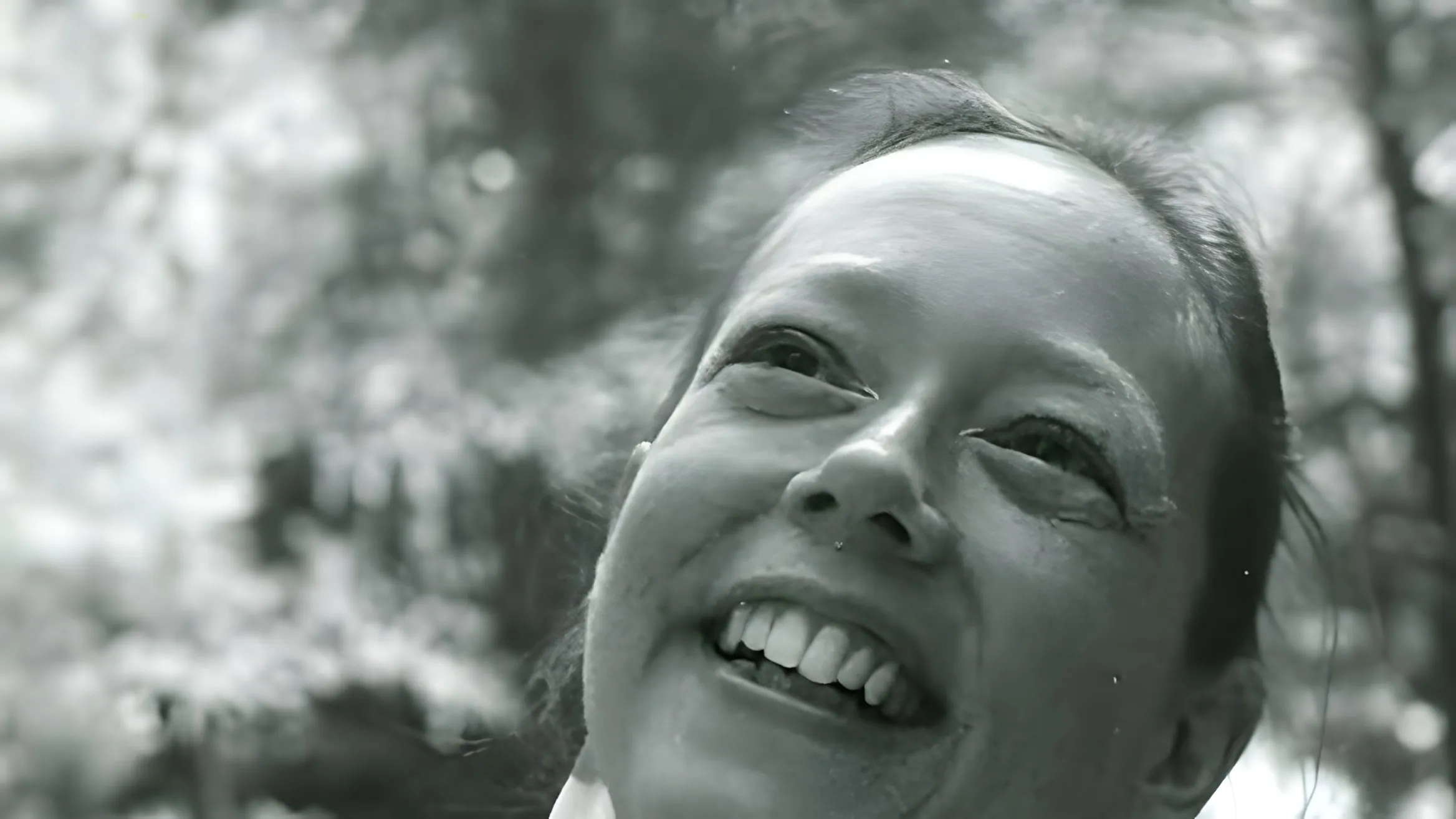 Ritratto in bianco e nero di Carla Rueckert, che sorride con il viso rivolto verso l’alto, all’aperto tra gli alberi