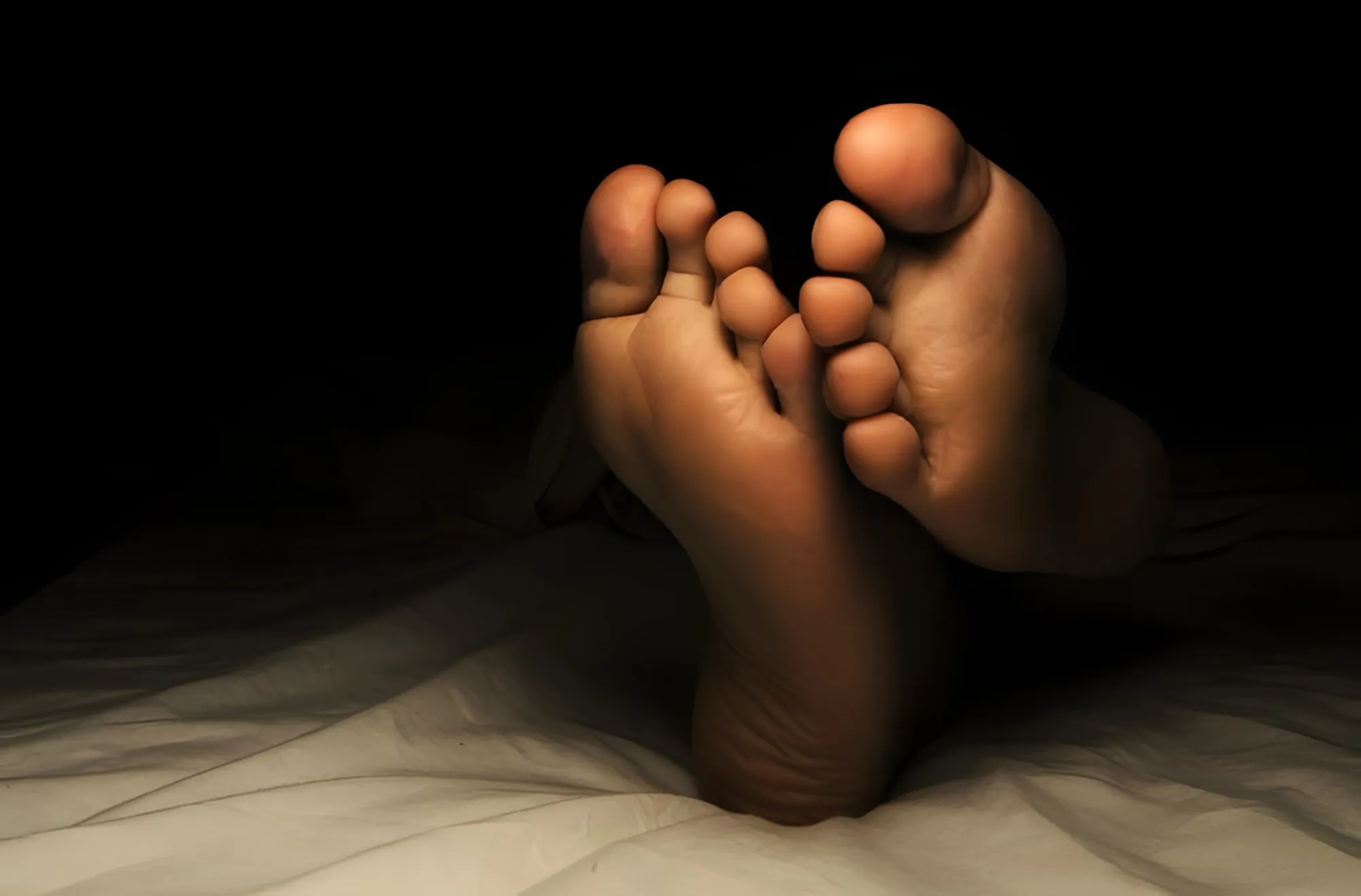 Piante dei piedi di una persona sdraiata nel buio, illuminate da una luce soffusa sulle lenzuola