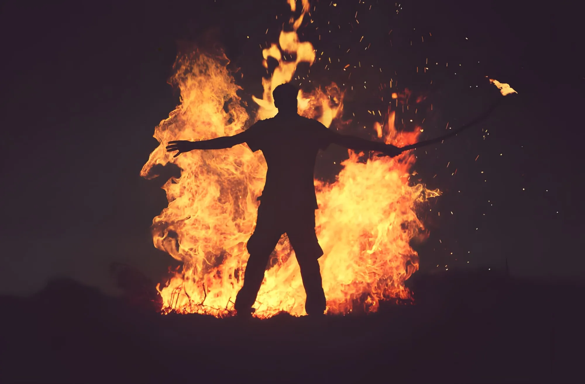 Sagoma di un uomo a braccia aperte davanti a grandi fiamme nella notte, con scintille nell'aria