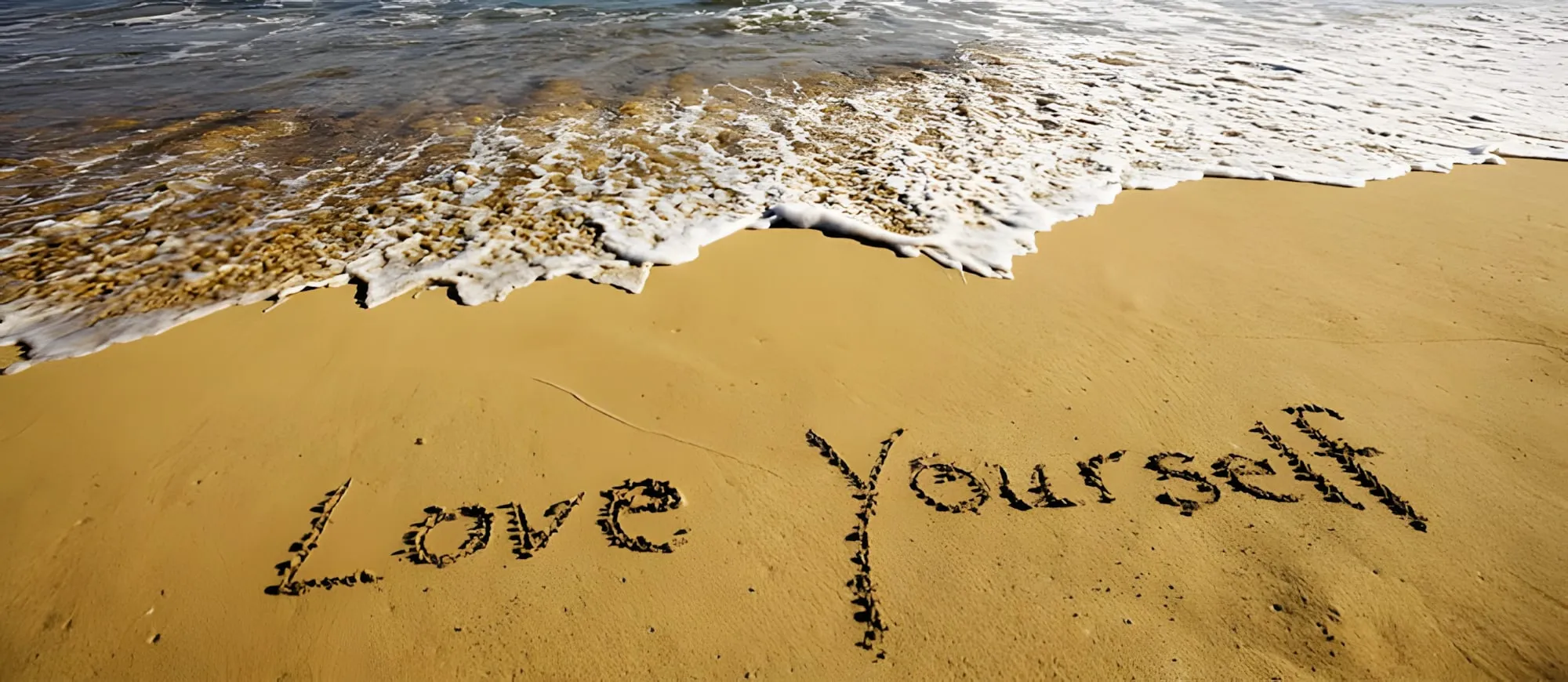 Scritta "Love Yourself" tracciata sulla sabbia dorata, vicino alla schiuma delle onde