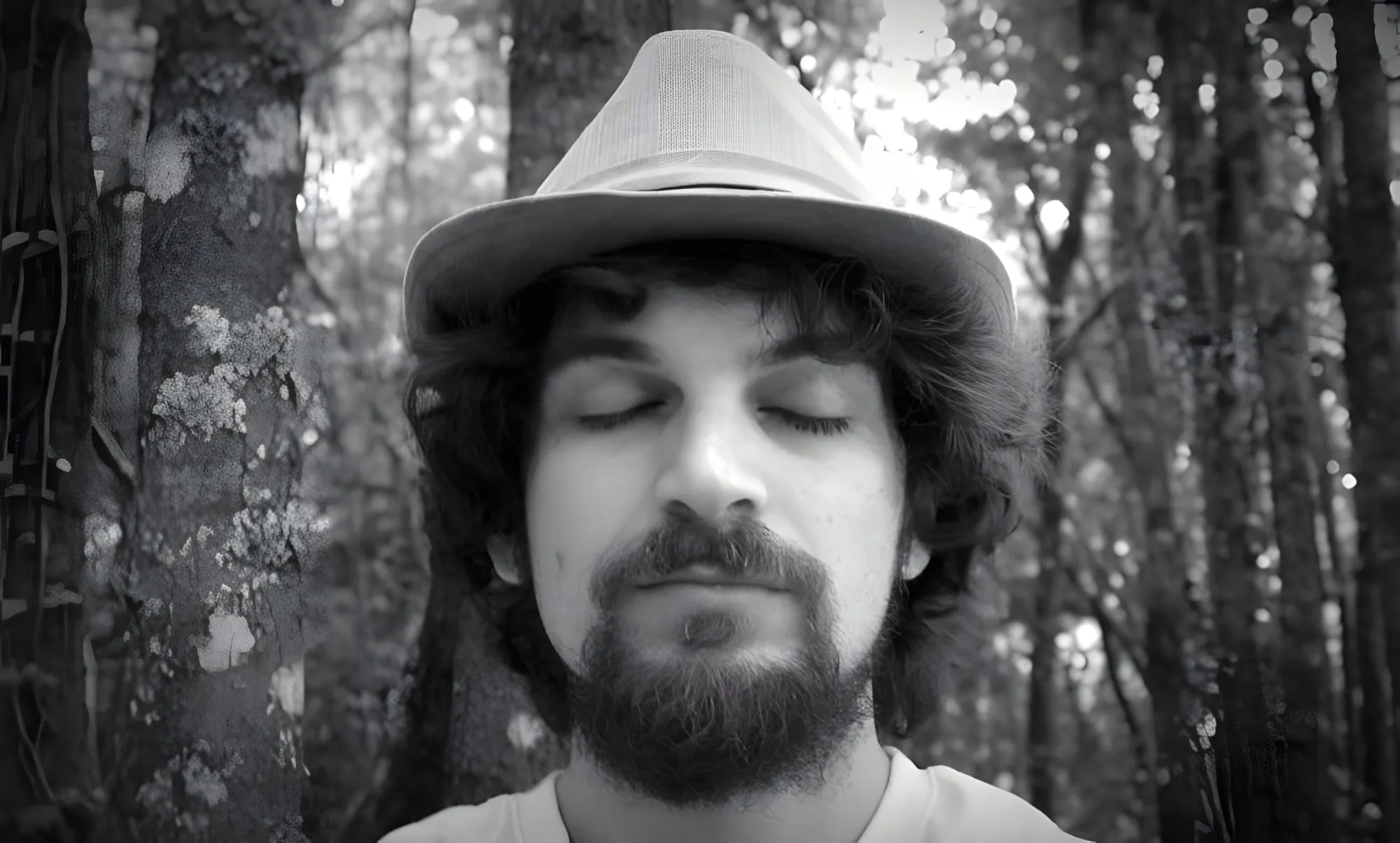 Ritratto in bianco e nero di un uomo con barba e cappello, a occhi chiusi, tra gli alberi di un bosco