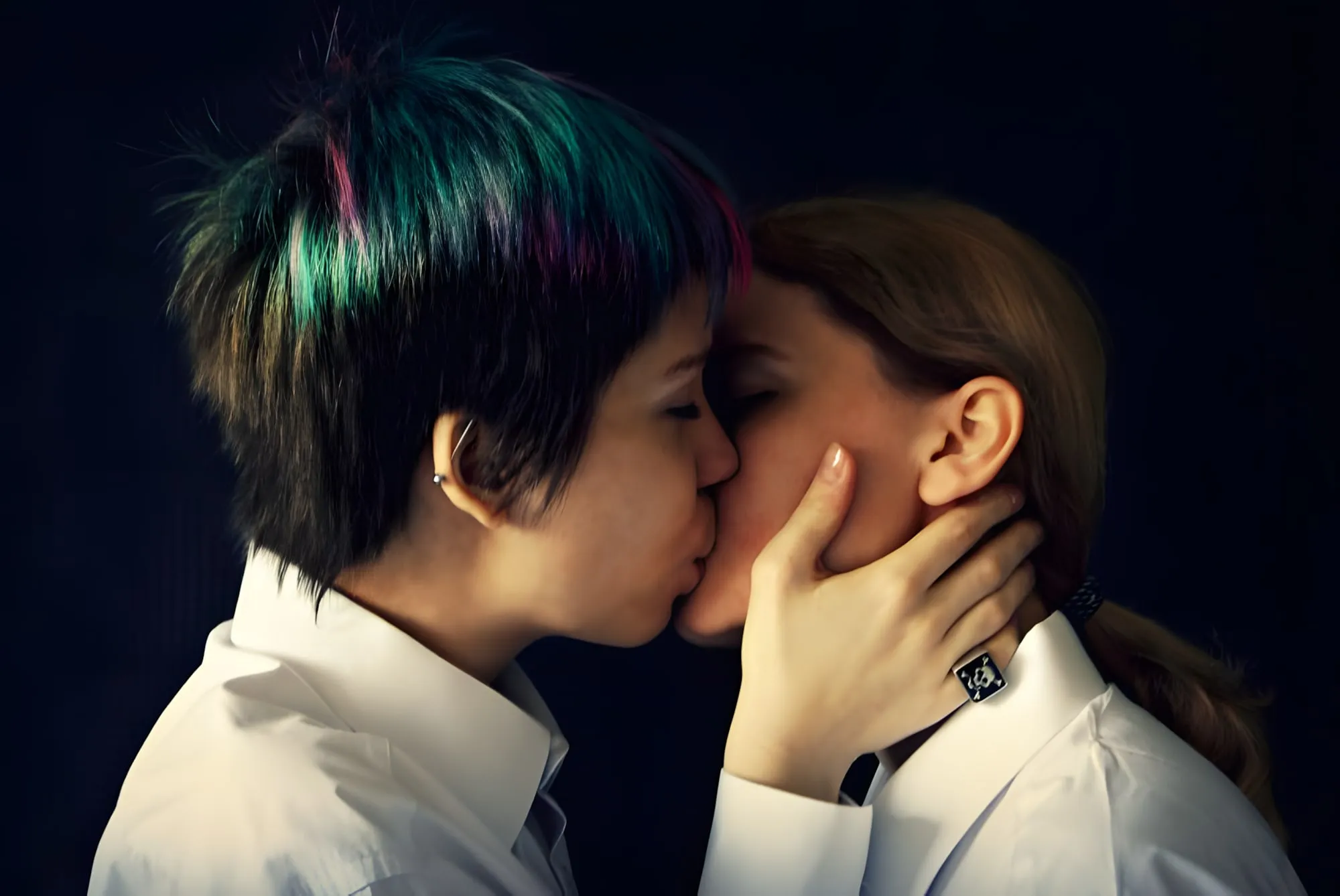 Due giovani donne che si baciano, una con i capelli corti e ciocche colorate, l’altra con i capelli raccolti, entrambe in camicia bianca