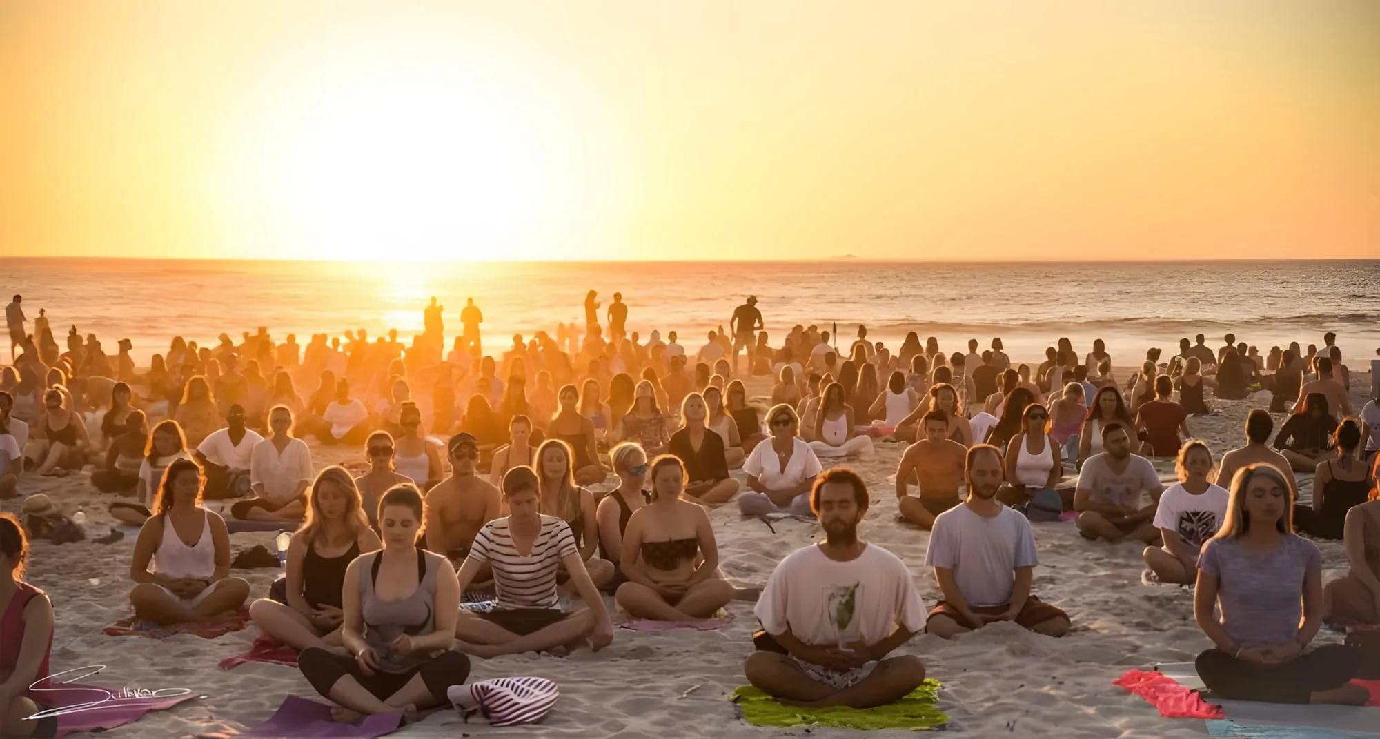 Centinaia di persone sedute in meditazione su una spiaggia al tramonto