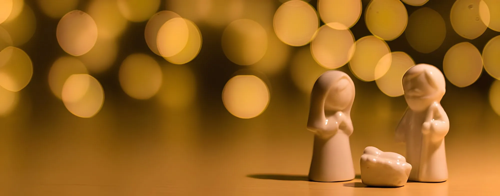 Tre statuine bianche di ceramica, la Madonna, San Giuseppe e il Bambino, davanti a luci dorate sfocate