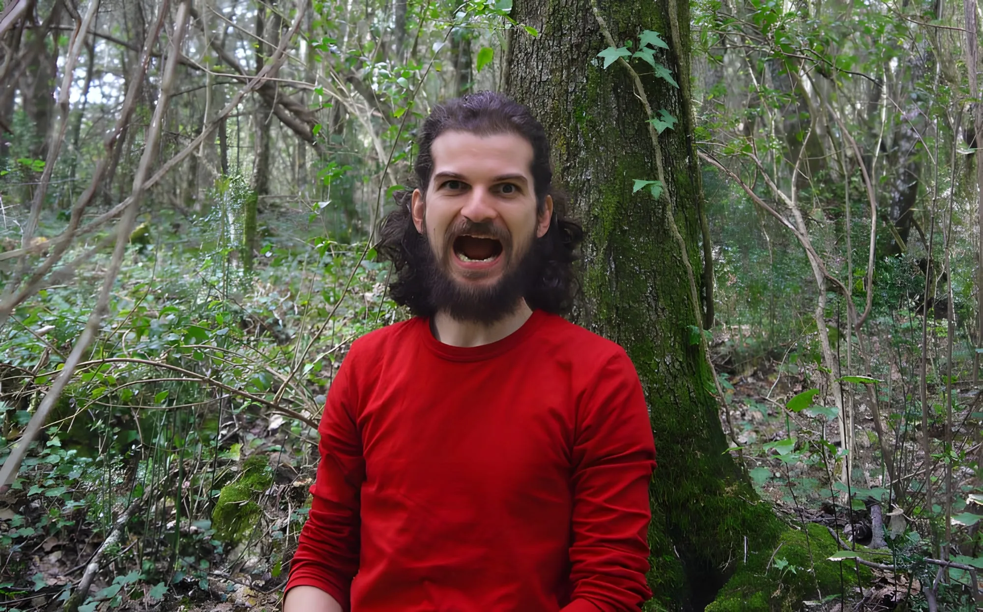 Uomo barbuto con maglia rossa che urla a bocca spalancata davanti a un tronco coperto di muschio in un bosco