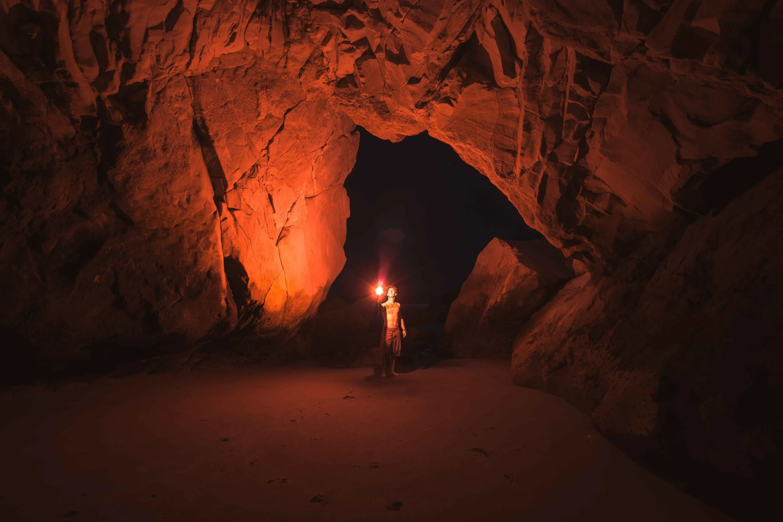 Uomo che tiene una torcia accesa in una grande caverna di roccia rossa, aperta sul buio della notte