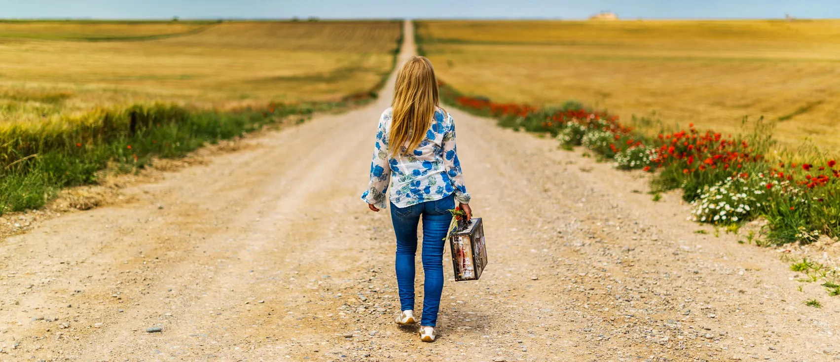 Una donna di spalle cammina con una valigetta lungo una strada sterrata tra campi di grano e papaveri