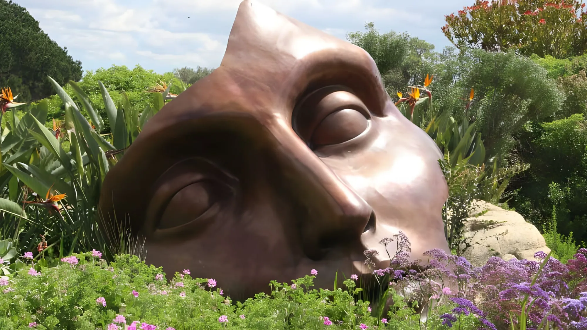 Grande scultura in bronzo raffigurante un volto, con gli occhi chiusi, adagiata in un giardino fiorito