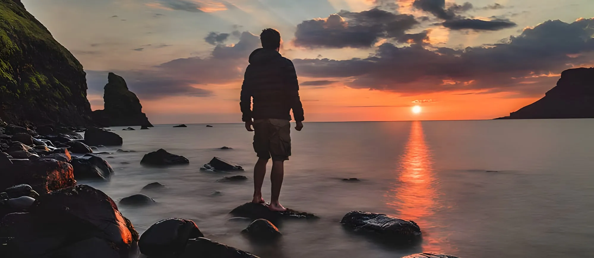 Uomo di spalle, a piedi nudi su uno scoglio, guarda il sole che tramonta sul mare